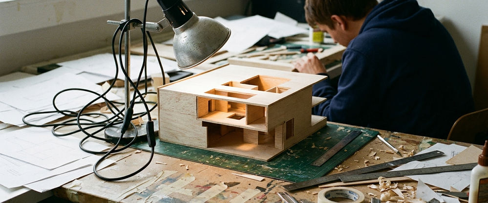 Photography setup with lighting equipment angled awkwardly toward a half-finished balsa architectural model